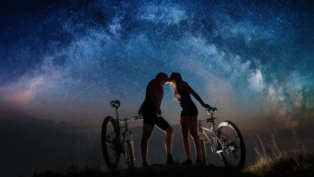 Nightly rearview of couple with mountain bikes kissing on a hill, watching night landscape with stars