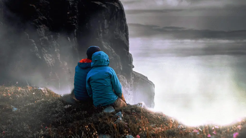 Couple in rain gear sitting on a windy, mossy rock looking out at the stormy sea