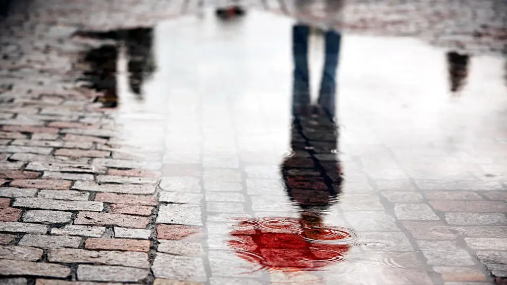 Reflection of young man with red umbrella in puddle on a rainy city street during rain.